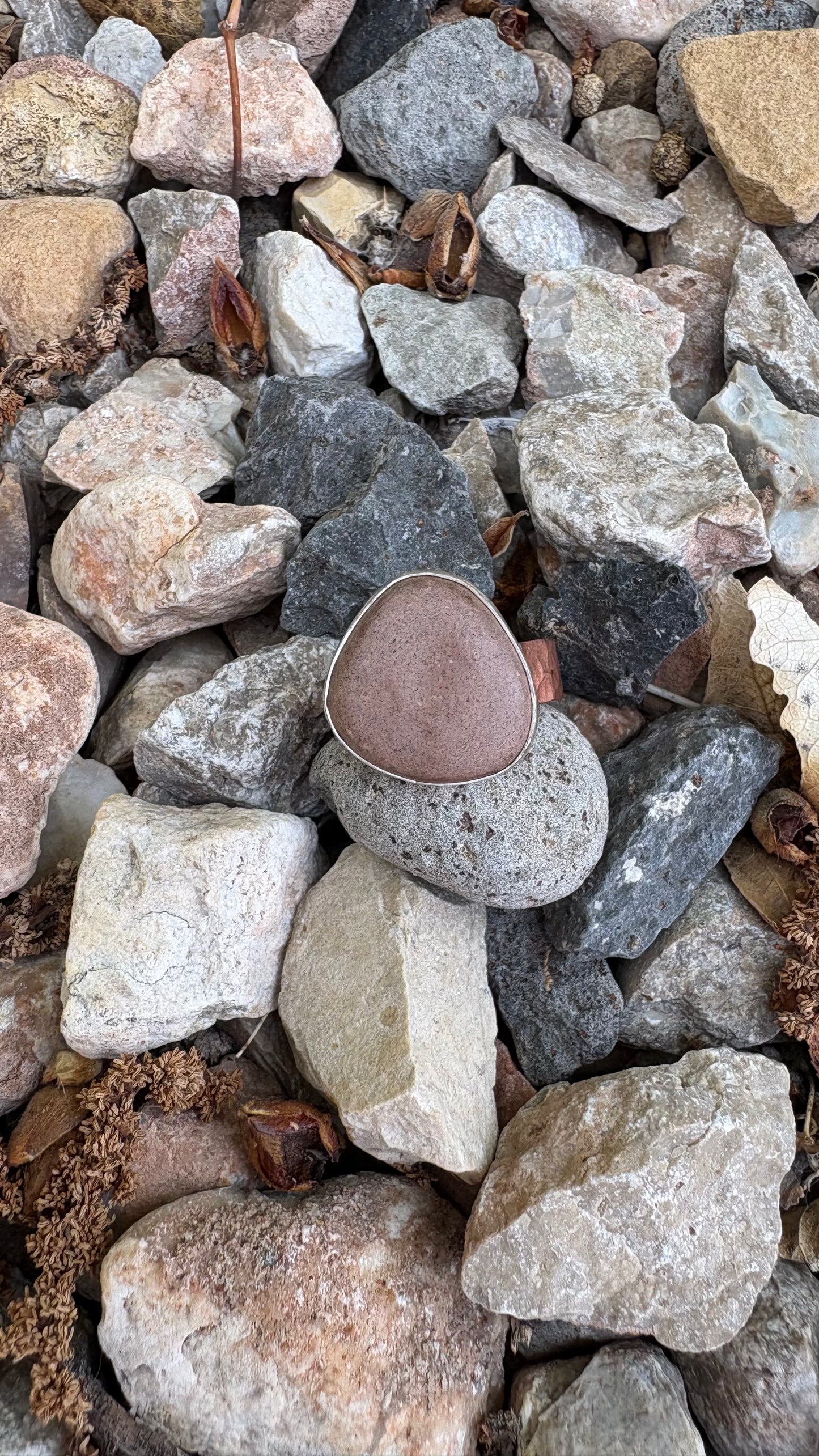 Close up of a silver and copper ring with reddish brown clay on a bed of multi colored rocks.