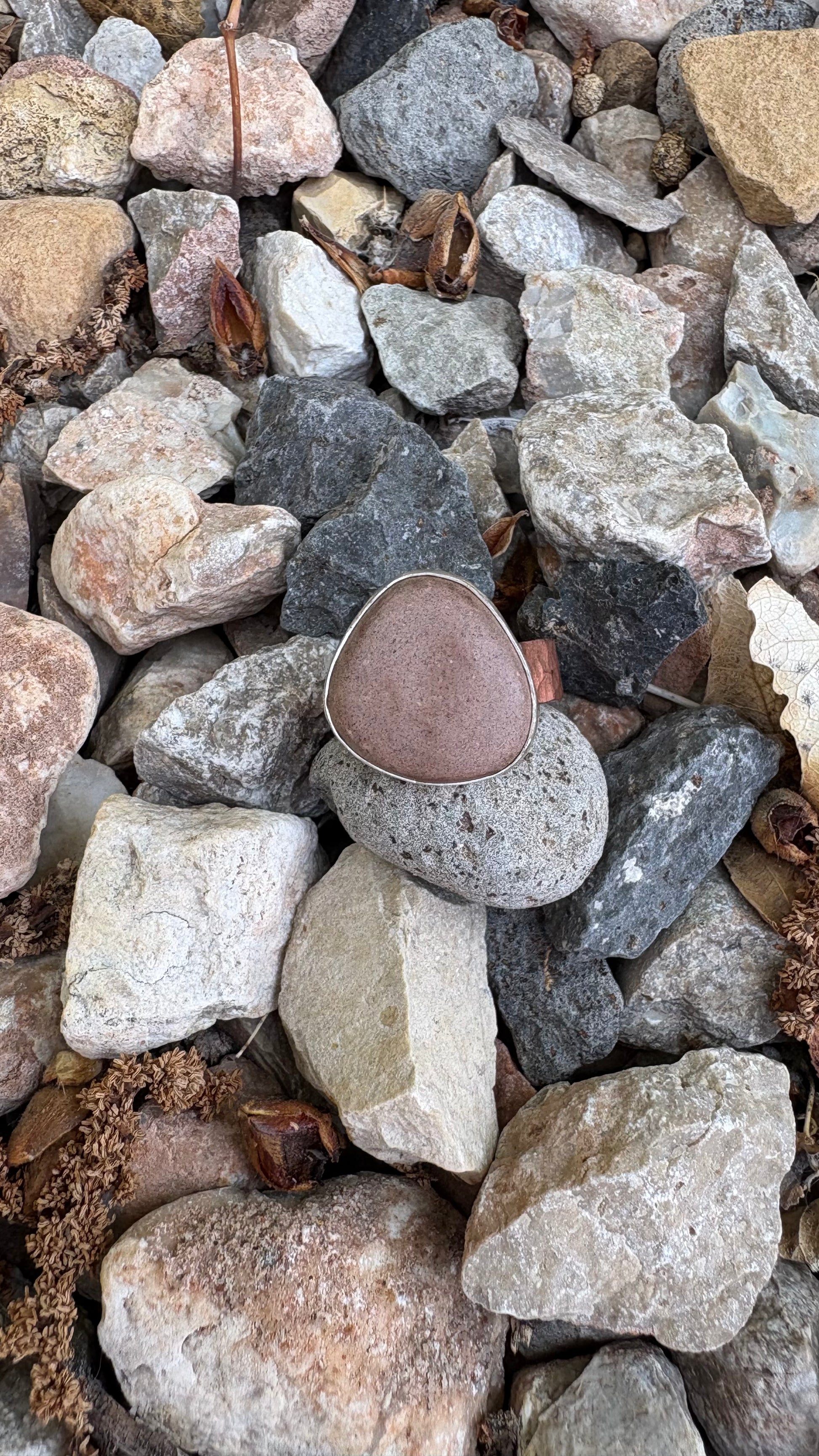 Close up of a silver and copper ring with reddish brown clay on a bed of multi colored rocks.