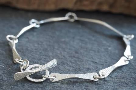A sterling silver bracelet with a hammered finish, featuring a toggle clasp and textured bar links, displayed on a dark slate surface.