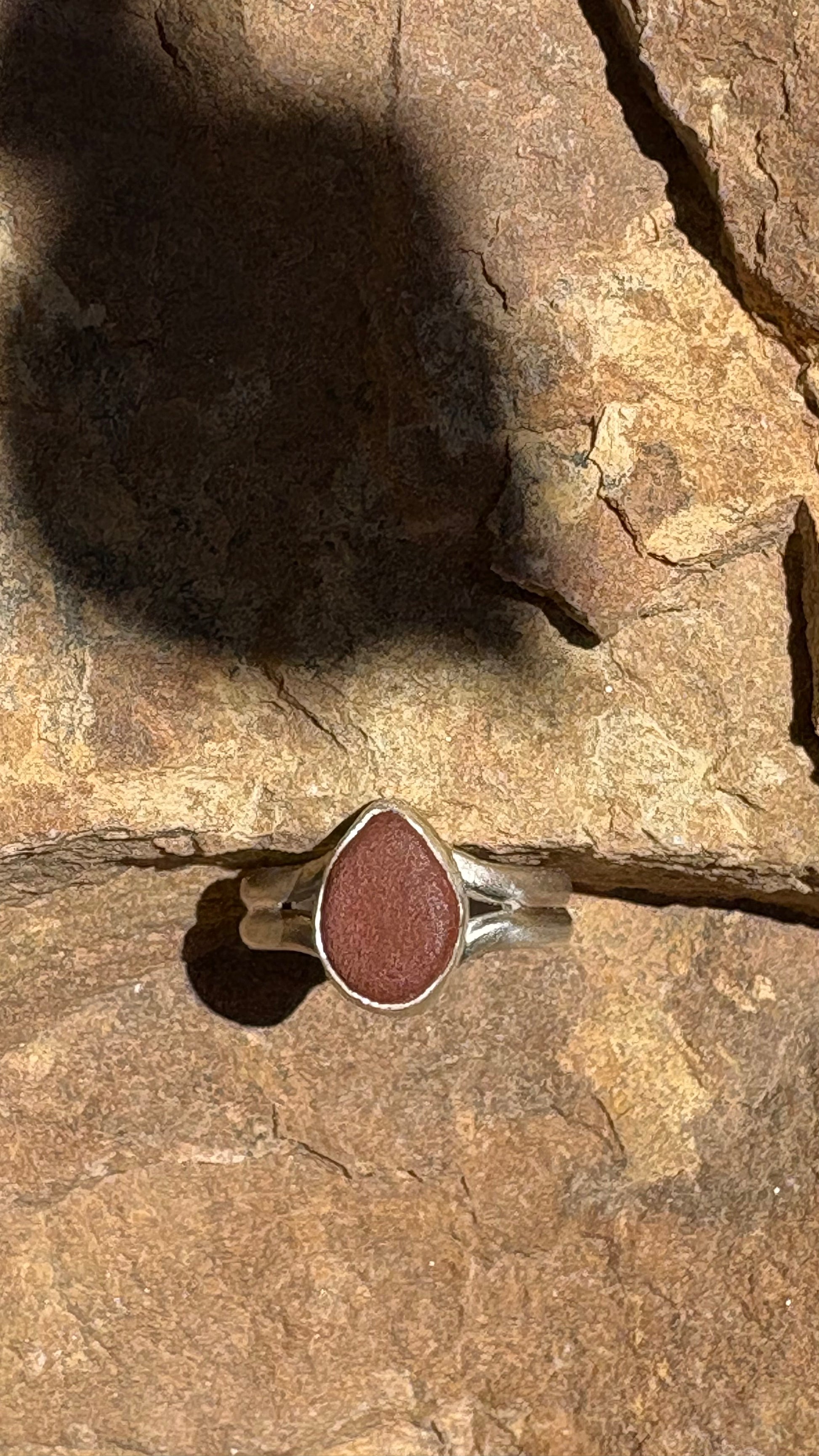 A silver ring with a teardrop-shaped reddish-brown stone, photographed resting on a textured brown rock surface under direct sunlight.