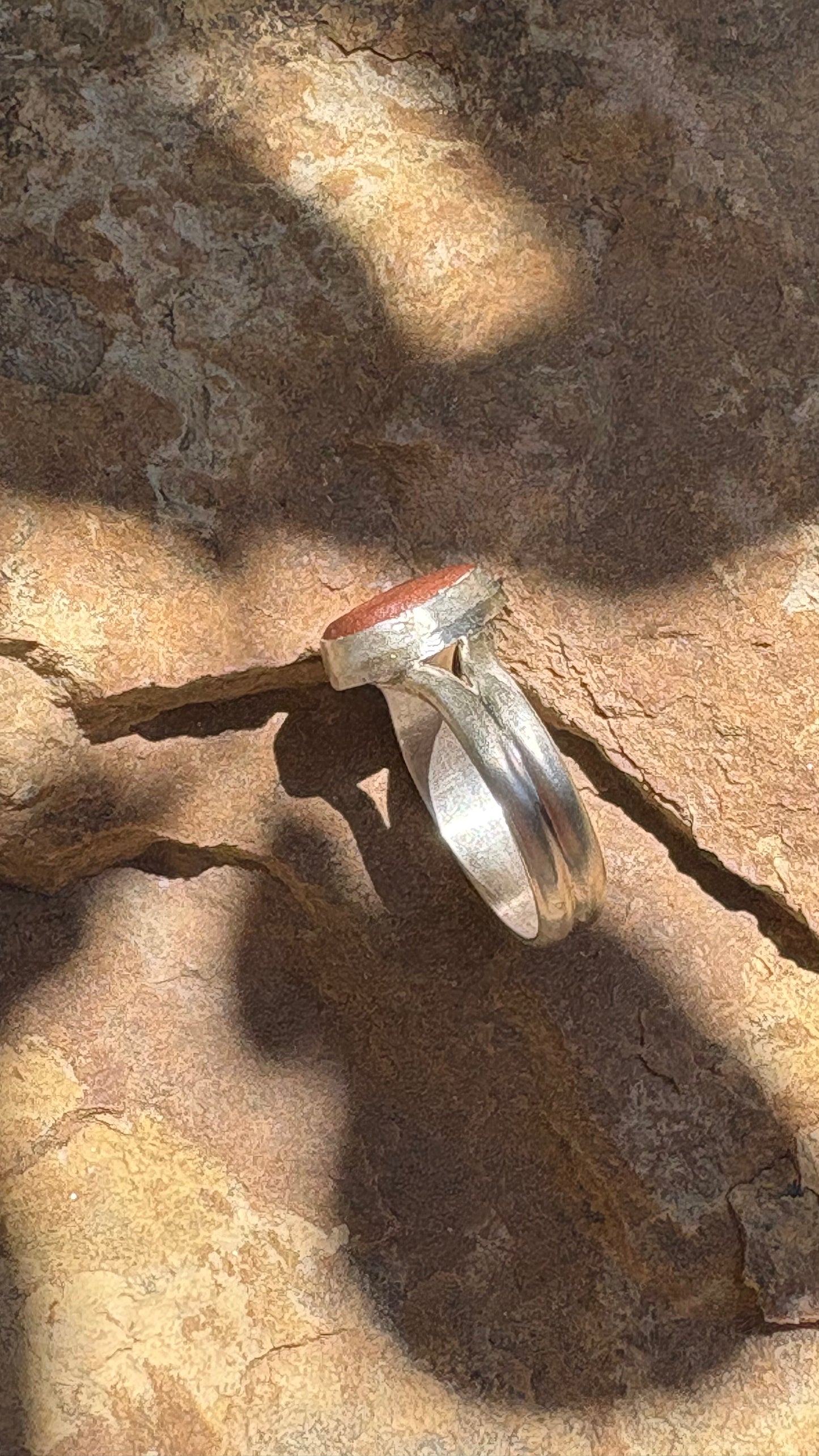A silver ring with a round reddish-brown stone, photographed resting on a textured brown rock surface under dappled sunlight and shadows.