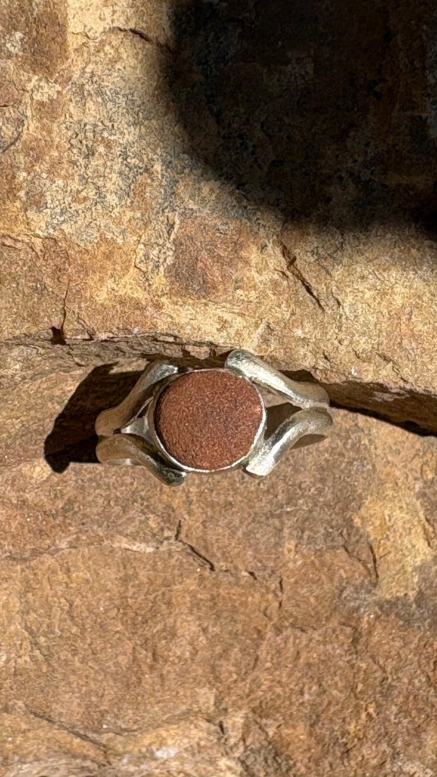 A silver ring with a round, textured reddish-brown stone, photographed resting on a textured brown rock surface.