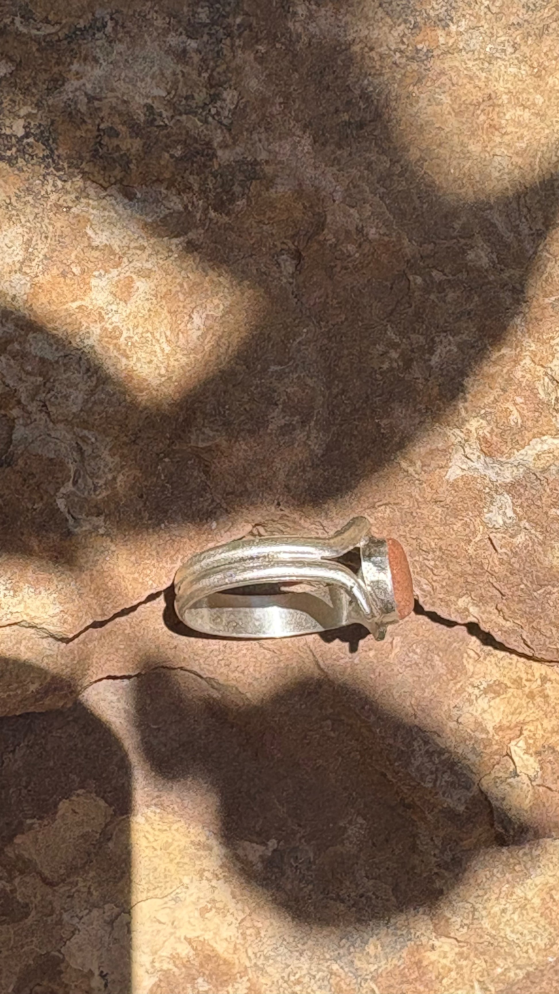 A silver-toned ring with a round orange stone, photographed on a textured brown rock surface with strong natural shadows.