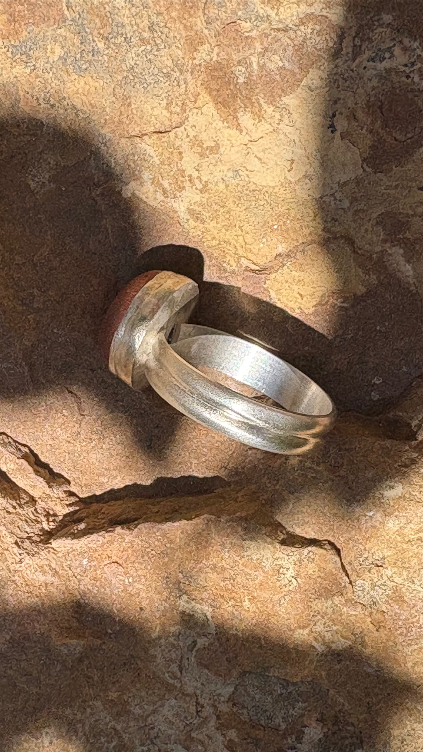 A silver-toned textured ring with a small reddish-brown accent on the side, resting on a textured brown rock surface under direct sunlight.