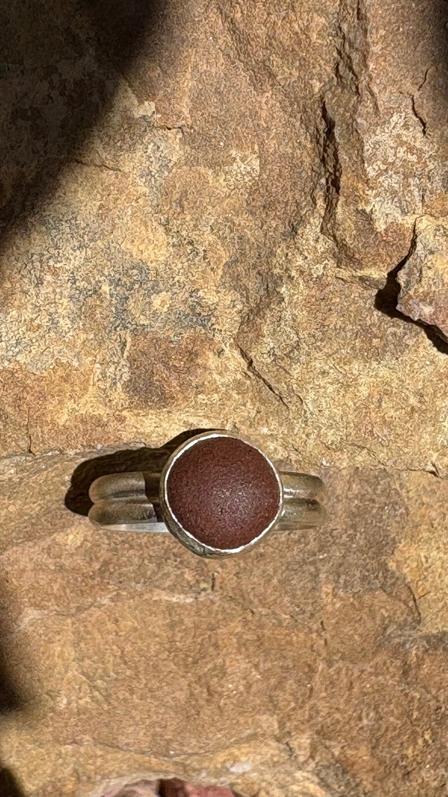 A silver band ring with a round, textured reddish-brown stone, photographed resting on a textured brown rock surface.