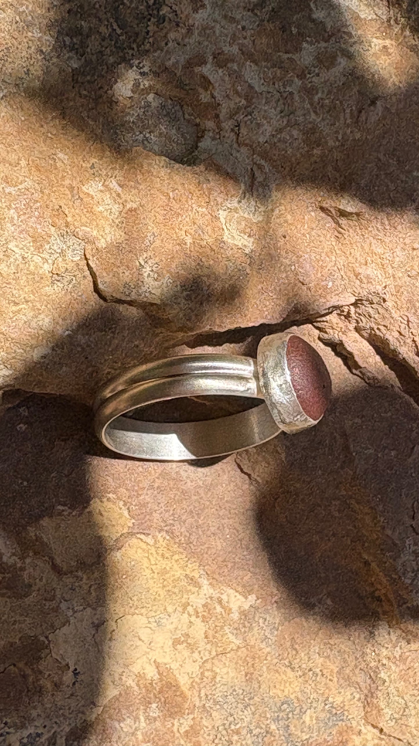 A silver-toned ring with a round reddish-brown stone, photographed resting on a textured rock surface in direct sunlight.