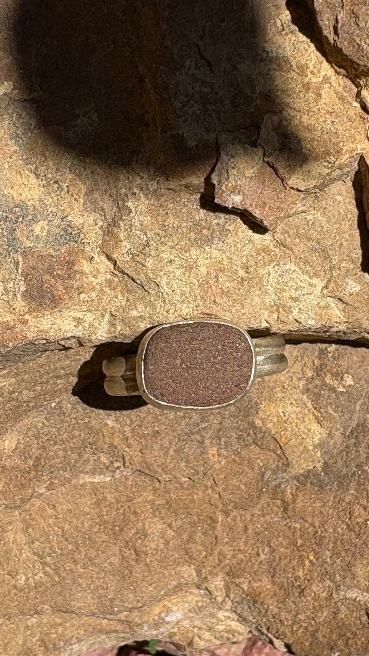 A silver-toned ring with a square cushion-cut brown textured stone, photographed lying on a textured brown rock surface in direct sunlight.