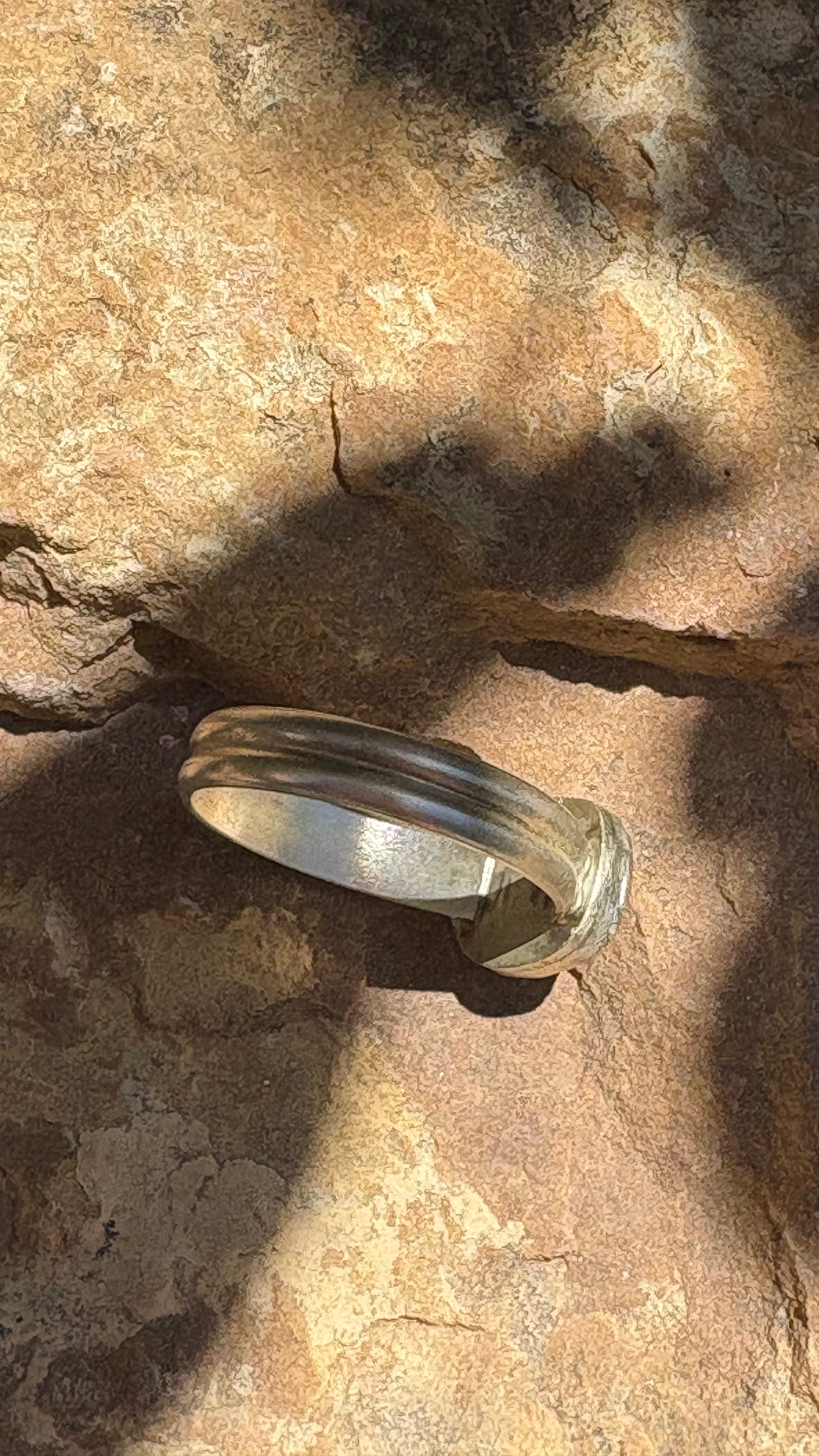 A polished silver-toned band ring resting on a textured brown rock surface under natural sunlight with shadows.